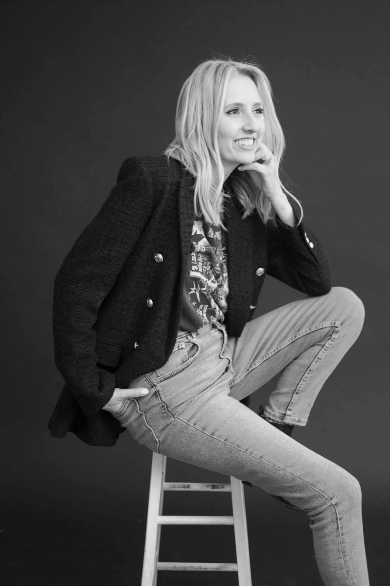 Kitty Dickerson-Firth seated on a wooden stool in a tweed blazer, graphic t-shirt and high-waisted jeans, photographed in black and white against a dark studio backdrop